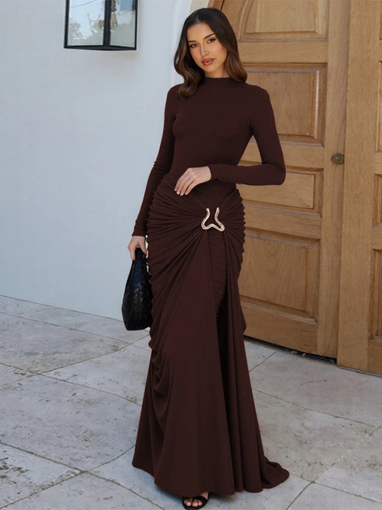 Woman wearing an auburn long-sleeve draped maxi dress with metal hip detail in a modern living room setting.