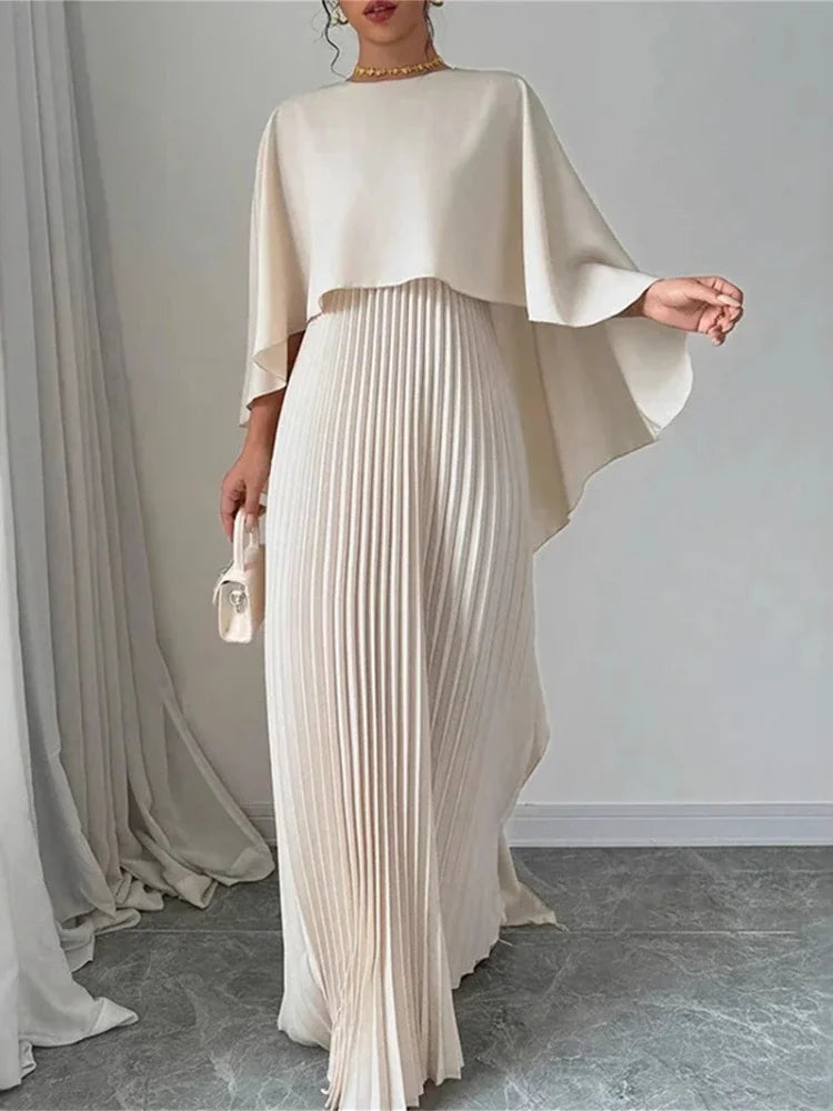 Woman wearing an ivory cape overlay pleated maxi dress in a minimalist setting.